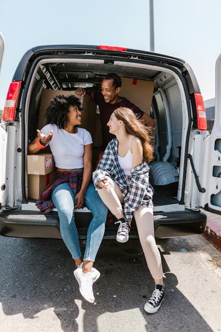 A man wearing a white polo shirt, dark cap, and jeans is standing inside the back of a moving van, holding a cardboard box with both hands during a home relocation. The van's interior is filled with n