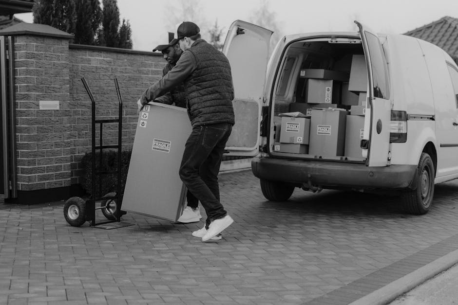 Two movers from Man with Van Wandsworth are engaged in loading cardboard boxes and furniture onto a small delivery van parked outside a residential property. One mover, wearing a cap and jacket, is guiding a large box onto a wheeled trolley, while the other is assisting with lifting and positioning the item. The back of the van is open, revealing an interior filled with packed boxes and household items prepared for home relocation. The scene takes place on a paved driveway adjacent to a brick wall and a residential building, with the movers using protective packing materials like plastic wraps or blankets on the furniture. The setting illustrates furniture transport and packing procedures typical of professional removals services along with careful handling during the loading process, associated with house moving near Wandsworth Common.