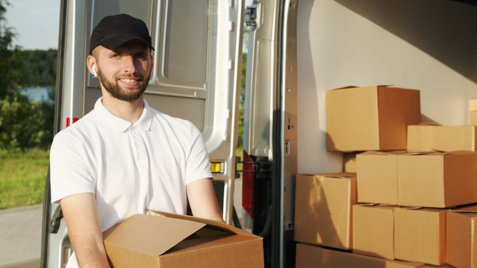 A man with a beard and wearing a black cap and white polo shirt stands outside the rear of a white moving van, holding a cardboard box during a home relocation process. Several packed cardboard boxes are stacked inside the van, ready for transportation, with some boxes placed on the pavement nearby. The van’s rear door is open, revealing a spacious interior with additional boxes and packaging materials, such as plastic wrap and protective blankets. The background features a partly cloudy sky and greenery, indicating a residential area near Wandsworth Common. The scene captures the loading process involved in furniture transport and packing for a house move, highlighting the professional services offered by Man with Van Wandsworth for removals and local moving logistics.